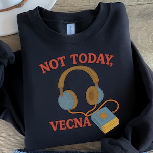 May include: A navy blue sweatshirt featuring the words "NOT TODAY, VECNA" in orange. The design includes brown headphones with blue ear pads and a small blue book. The sweatshirt is folded.