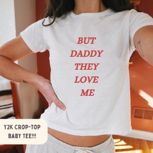 May include: White crop top with the text "BUT DADDY THEY LOVE ME" in red lettering. The text is centered on the shirt. The shirt is a Y2K style crop top.