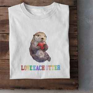 May include: White t-shirt featuring a cartoon illustration of a sea otter holding a red heart. The text "LOVE EACH OTTER" is printed below the otter in rainbow colours.