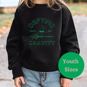 May include: Black sweatshirt with green text that reads "Defying Gravity" with a witch hat and broom graphic. The text also says "Since 2001". The sweatshirt is available in youth sizes.