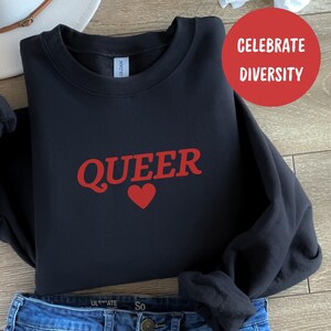 Op de afbeelding: Zwart sweatshirt met het woord "QUEER" in rode letters en een rood hart. De tekst "VIER DE DIVERSITEIT" staat in rood op een witte cirkel.