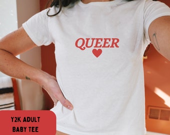 Może przedstawiać: Biała, krótka koszulka z napisem "QUEER" w czerwonych literach i czerwonym sercem. Koszulka jest oznaczona jako "Y2K ADULT BABY TEE".