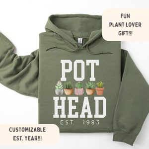Pode incluir: Camisola de capuz verde com o texto "POT HEAD" em branco, com uma fila de plantas em vasos abaixo do texto e "EST. 1983" abaixo das plantas.
