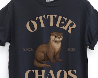 Puede incluir: Camiseta azul marino con una ilustración de nutria marrón. El texto "OTTER CHAOS" está arqueado sobre la nutria, con "SINCE 2025" debajo. La camiseta está hecha de un material suave y cómodo.