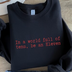 Op de afbeelding: Zwart sweatshirt met de rode tekst: "In a world full of tens, be an Eleven". Het Gildan-merklabel is zichtbaar. Het gevouwen kledingstuk suggereert casual kleding met een inspirerende boodschap.