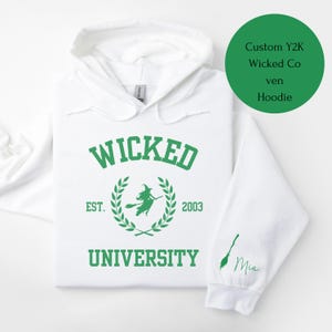 May include: White hooded sweatshirt with green text that reads "Wicked University" with a witch silhouette and laurel wreath. The text "Est. 2003" is below the wreath. The sleeve has a green broom and the name "Mia" written in cursive.