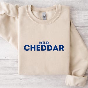 Puede incluir: Una sudadera beige con el texto "MILD CHEDDAR" impreso en azul.