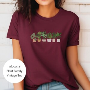 May include: A maroon t-shirt with a graphic of a row of potted plants. The text "Alocasia Plant Family Vintage Tee" is on a white circle in the lower left corner of the image.