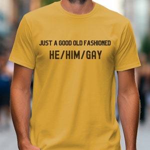Op de afbeelding: Een geel T-shirt met de tekst "JUST A GOOD OLD FASHIONED HE/HIM/GAY" gedrukt op de voorkant.