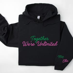 Könnte beinhalten: Schwarzer Kapuzenpullover mit dem Text "Together We're Unlimited" in grüner und rosa Schrift. Der Ärmel hat den Text "Mia + Ella" in rosa Schrift.