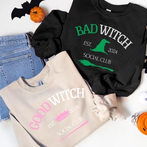 Puede incluir: Dos sudaderas con temática de Halloween. Una es beige con texto rosa que dice "Good Witch Est. 2024 Social Club" y una corona y una varita rosas. La otra es negra con texto verde que dice "Bad Witch Est. 2024 Social Club" y un sombrero de bruja verde y una escoba.