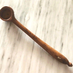 May include: A long-handled brown wooden spoon with a small, deep, circular bowl at one end and a tapered handle that narrows toward the tip. The wood features a natural grain pattern and a small white mark near the end of the handle.