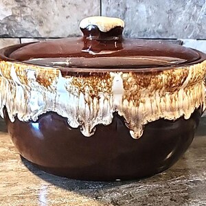 May include: A round ceramic casserole dish with a lid, featuring a glossy dark brown base and a decorative cream-coloured drip glaze pattern around the upper rim and the lid handle. The pot includes two small side handles for lifting.
