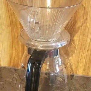 Peut inclure: Un cône de filtration à café en verre transparent avec des rainures intérieures verticales repose sur une carafe en verre transparent. La carafe est dotée d'une poignée en plastique noir et d'un large rebord circulaire, révélant sa structure.