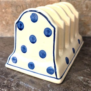 Puede incluir: Un soporte para tostadas de cerámica color crema con un patrón de lunares azules pintados a mano. La pieza tiene una forma de arco redondeado con cuatro ranuras verticales para sostener rebanadas de pan, con un acabado brillante y bordes azules.