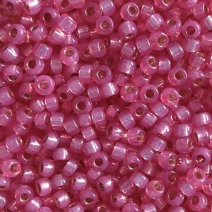 Pode incluir: Uma coleção de pequenas contas de vidro rosa translúcido, redondas, com um acabamento brilhante e iridescente. Cada conta apresenta um orifício central, criando uma aparência uniforme em toda a superfície. As contas estão amontoadas.