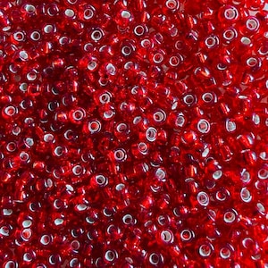 Puede incluir: Una colección de pequeñas cuentas de vidrio rojo translúcido con forma circular y un orificio central. Las cuentas están densamente agrupadas, creando una textura uniforme de tonos rojos vibrantes con reflejos de luz visibles en las superficies.