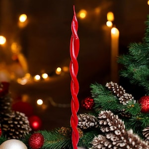 Può includere: Una candela rossa attorcigliata con un tema natalizio festoso. La candela è circondata da rami di pino, pigne e decorazioni rosse.