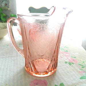 May include: A translucent pink glass pitcher featuring an intricate embossed floral pattern on its exterior. The vessel includes a curved handle on one side, a wide pouring spout at the top, and a circular base, showcasing a delicate pressed glass design.