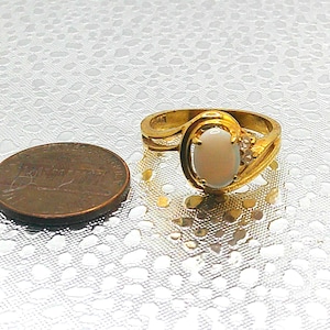 May include: A gold-toned ring with an oval, light-colored stone. The ring has a decorative swirl design and small accent stones. A penny is next to the ring for size comparison.