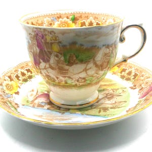 May include: A vintage teacup and saucer set. The cup and saucer are cream-colored with a detailed pastoral scene in shades of brown, green, and yellow. The rims are trimmed with gold. The cup has a curved handle.