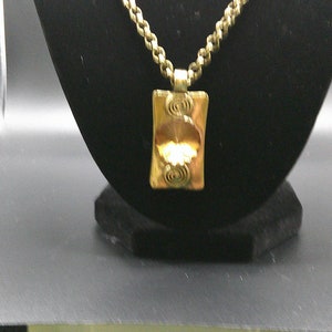 May include: Gold-toned pendant necklace. The rectangular pendant features a central design with a floral element and spiral accents. The necklace has a textured chain and is displayed on a black stand.