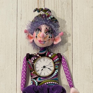 May include: A whimsical doll with a clock face, featuring a purple ruffled skirt and striped blue legs. The doll has a patterned bodice, purple polka dot sleeves, and a matching hat. The doll's face is detailed with painted features and pink accents.