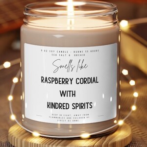 May include: A 9 oz soy candle in a clear glass jar with a white label. The label reads "Smells like Raspberry Cordial with Kindred Spirits". The candle is lit and the flame is visible. The candle is sitting on a wooden coaster.