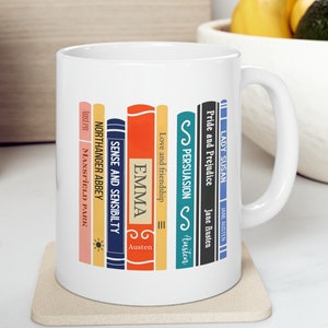 May include: White ceramic mug featuring a colourful design of stacked books with the titles of Jane Austen novels. The titles include "Pride and Prejudice", "Persuasion", "Emma", "Sense and Sensibility", "Northanger Abbey", and "Mansfield Park".