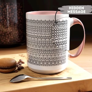 May include: White ceramic mug with a pink interior and handle, adorned with a black geometric pattern. A silver spoon and a macaron rest on a wooden board. The text "HIDDEN MESSAGE" is visible.