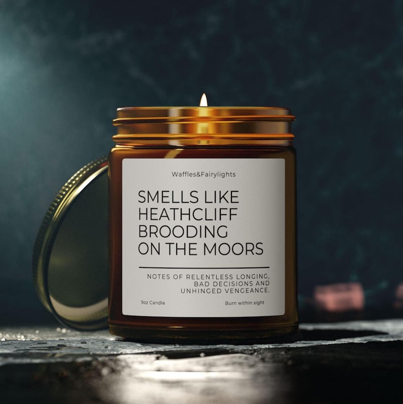 May include: A lit candle in a brown glass jar with a gold lid. The label reads "SMELLS LIKE HEATHCLIFF BROODING ON THE MOORS." The candle is 9 oz and has a scent described as "NOTES OF RELENTLESS LONGING, BAD DECISIONS AND UNHINGED VENGEANCE."