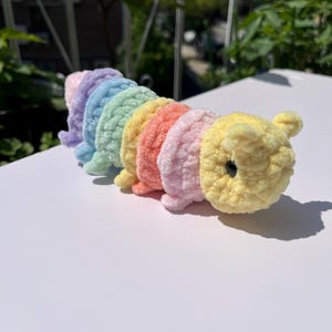 May include: A handmade, plush caterpillar toy in a rainbow of colors: lavender, blue, green, yellow, orange, and pink. The caterpillar has a yellow head with small ears and a black eye. The toy is made of soft, textured yarn.