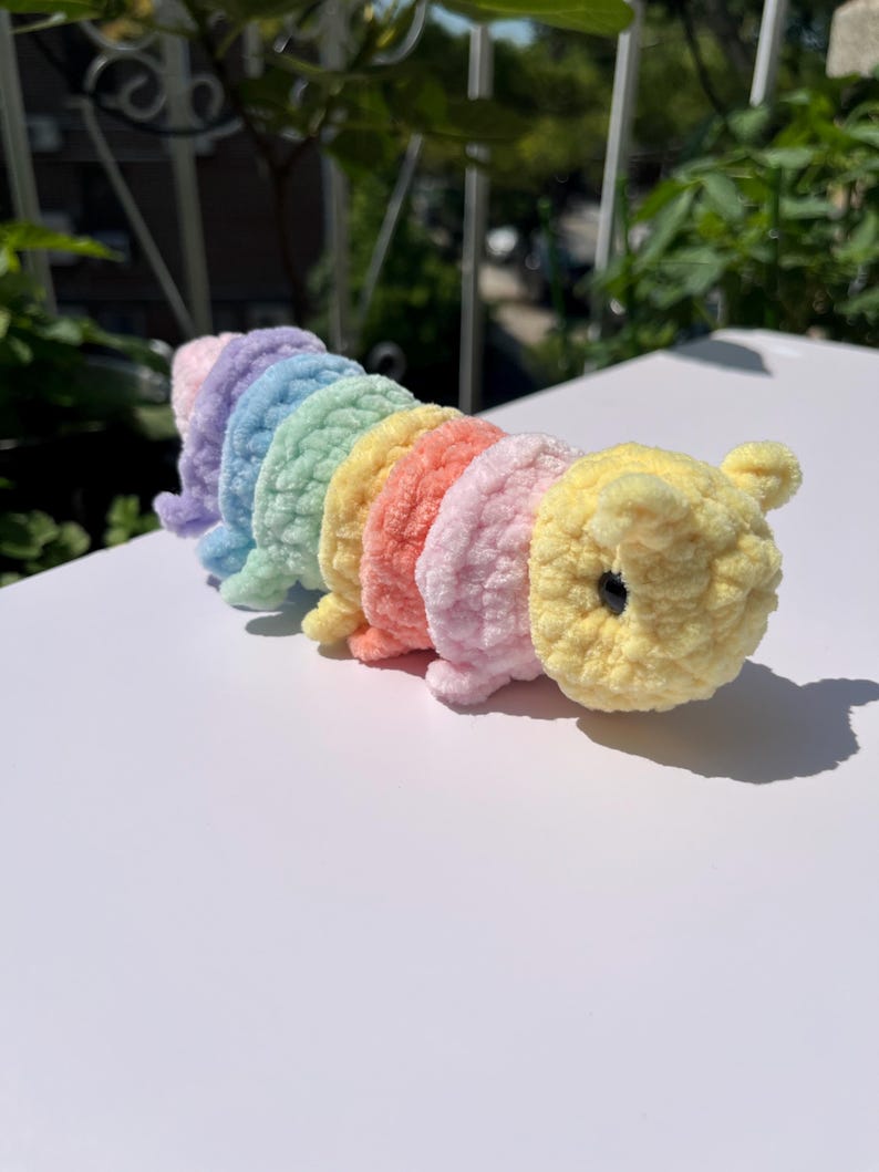 May include: A handmade, plush caterpillar toy in a rainbow of colors: lavender, blue, green, yellow, orange, and pink. The caterpillar has a yellow head with small ears and a black eye. The toy is made of soft, textured yarn.