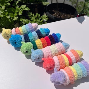 May include: A collection of colorful, crocheted caterpillar toys. Each caterpillar features a different color scheme, including yellow, blue, green, red, and purple. The toys are arranged on a white surface, with greenery in the background.