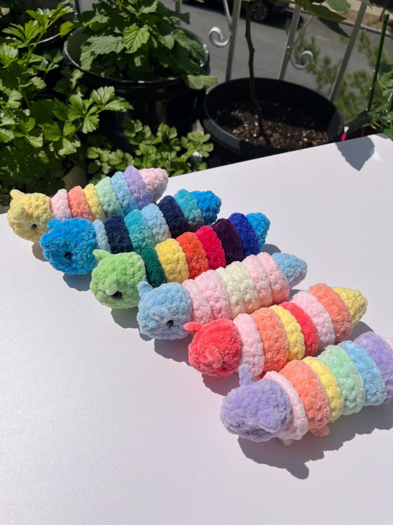May include: A collection of colorful, crocheted caterpillar toys. Each caterpillar features a different color scheme, including yellow, blue, green, red, and purple. The toys are arranged on a white surface, with greenery in the background.