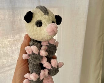 May include: A crocheted plush toy resembling a sugar glider. The toy is white with gray accents and pink feet and tail. The toy has black eyes and black ears.