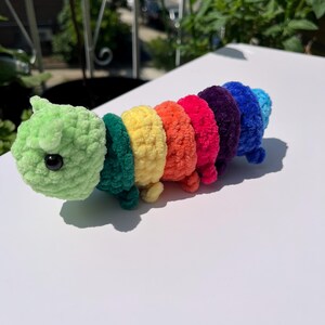 May include: A handmade, colorful crochet caterpillar toy. The caterpillar is made of soft, textured yarn in a rainbow of colors, including green, teal, yellow, orange, red, purple, and blue. It has a green head with a black eye and small legs.