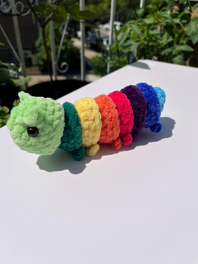 May include: A handmade, colorful crochet caterpillar toy. The caterpillar is made of soft, textured yarn in a rainbow of colors, including green, teal, yellow, orange, red, purple, and blue. It has a green head with a black eye and small legs.