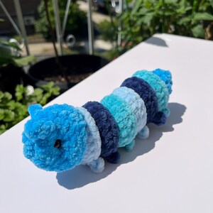 May include: A handmade crochet caterpillar toy in shades of blue. The plush toy features segments of varying blue hues, from light turquoise to deep navy. It has small, rounded legs and a small black bead for an eye. The caterpillar is displayed on a white surface.