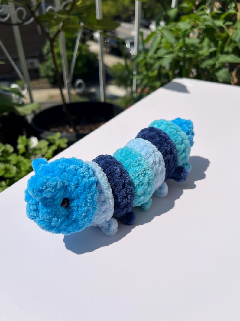 May include: A handmade crochet caterpillar toy in shades of blue. The plush toy features segments of varying blue hues, from light turquoise to deep navy. It has small, rounded legs and a small black bead for an eye. The caterpillar is displayed on a white surface.