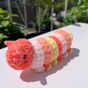 May include: A handmade, plush caterpillar toy in shades of pink, coral, yellow, and orange. The caterpillar has a segmented body, small legs, and a detailed head with tiny black eyes and antennae. The toy is made of soft, textured yarn.