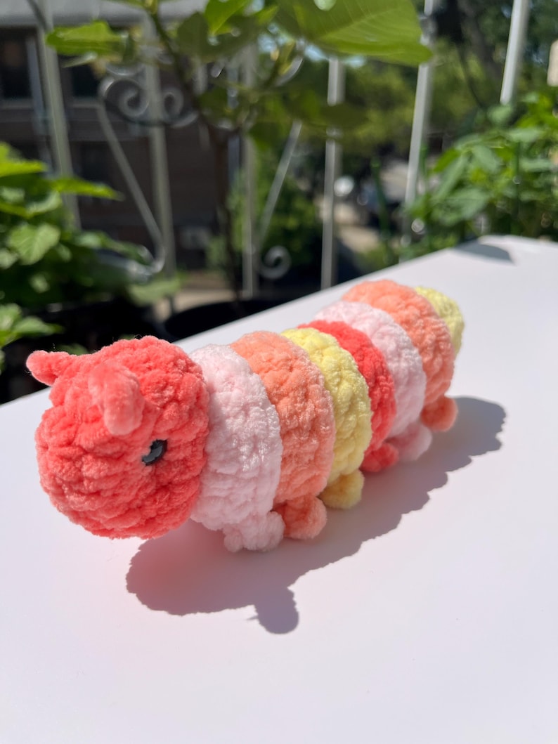 May include: A handmade, plush caterpillar toy in shades of pink, coral, yellow, and orange. The caterpillar has a segmented body, small legs, and a detailed head with tiny black eyes and antennae. The toy is made of soft, textured yarn.
