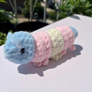 May include: A handmade, plush caterpillar toy in pastel colors. The caterpillar is made of soft, textured yarn in segments of light blue, pink, and cream. It has small, stubby legs and a round head with black eyes. The toy is approximately 6 inches long.