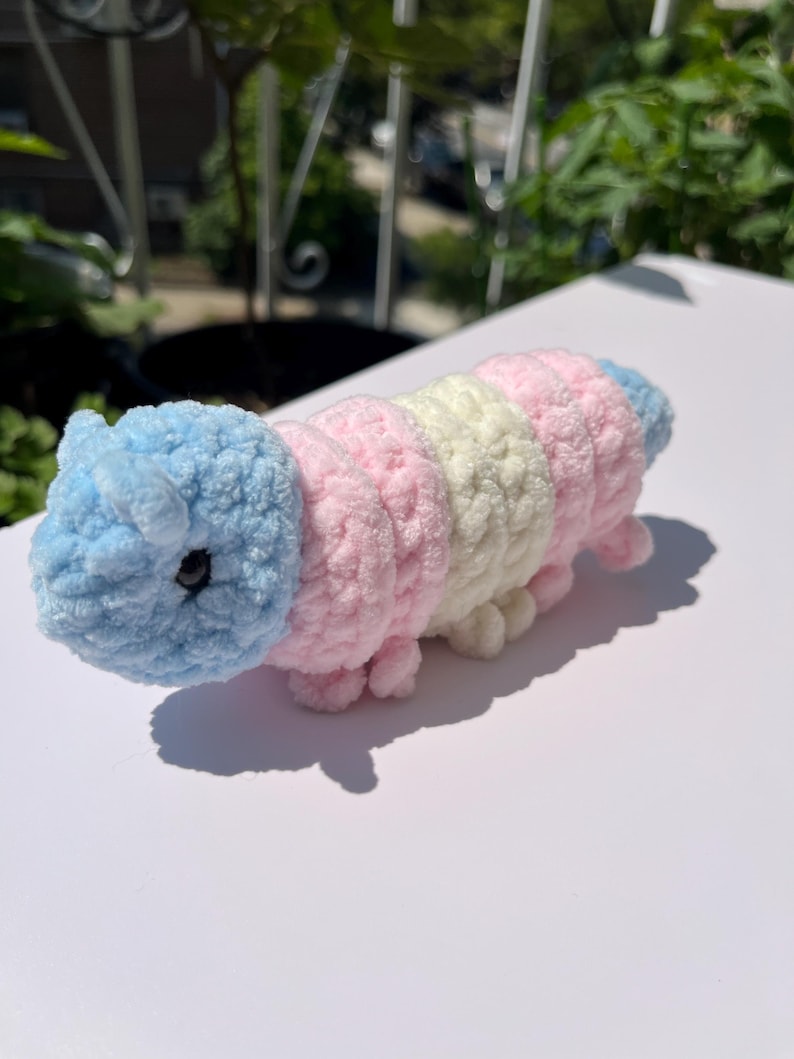 May include: A handmade, plush caterpillar toy in pastel colors. The caterpillar is made of soft, textured yarn in segments of light blue, pink, and cream. It has small, stubby legs and a round head with black eyes. The toy is approximately 6 inches long.