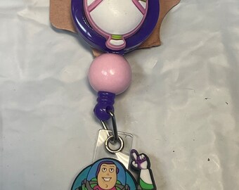 Puede incluir: Un carrete retráctil para placa con un diseño de dibujos animados de Buzz Lightyear. El carrete tiene un diseño de oreja de Mickey Mouse rosa y morado en la parte superior. El personaje de Buzz Lightyear está en un marco circular.