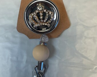 May include: A silver retractable badge reel with a white bead and a silver and gold crown charm with rhinestones.