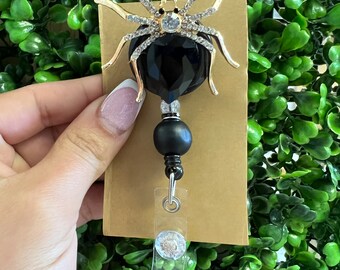 May include: Black spider-shaped badge holder with a black crystal and gold accents. The holder is attached to a black bead and a clear retractable badge reel.