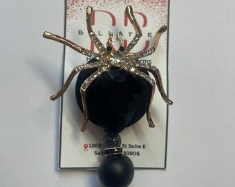 May include: A black and gold spider charm with rhinestones on a black retractable badge reel. The charm is attached to a black bead and a clear plastic badge clip.