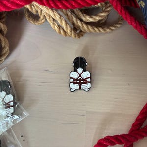 May include: A white and black enamel pin featuring a figure bound with red rope. The pin is set against a wooden surface, with red and tan ropes in the background. Additional pins are visible in a plastic bag.