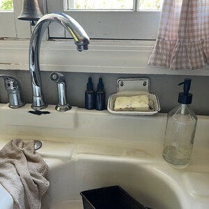 K&ouml;nnte beinhalten: Ein K&uuml;chenbecken mit einer verchromten Armatur, einer Seifenschale und zwei dunklen Glasflaschen. Ein klarer Glas-Seifenspender und zwei schwarze Brotformen befinden sich im Waschbecken. Ein beiges Handtuch h&auml;ngt an der Seite des Waschbeckens.